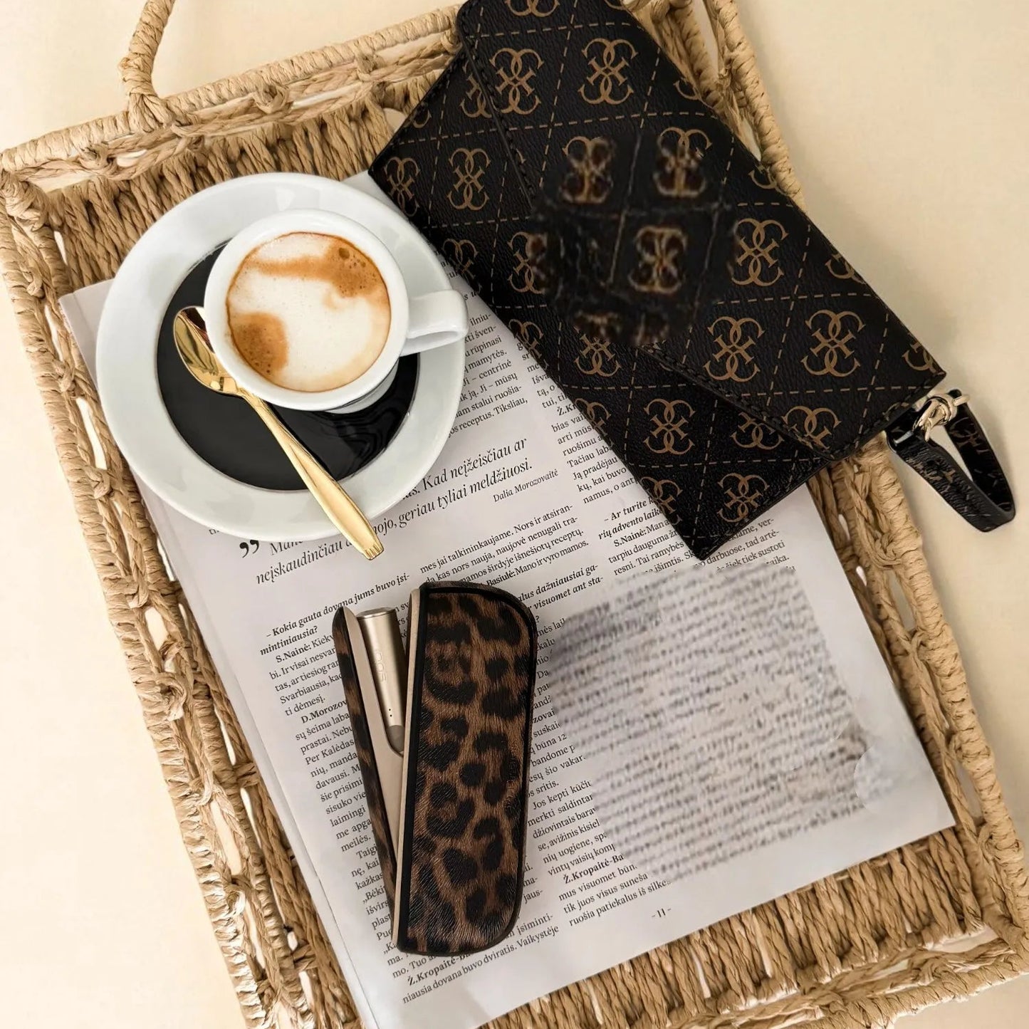 Cappuccino in a white cup with a gold spoon on a newspaper, with leopard print sunglasses and a patterned napkin on a woven tray.