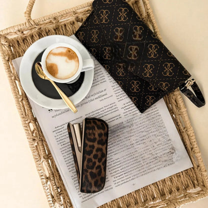 Cappuccino in a white cup with a gold spoon on a newspaper, with leopard print sunglasses and a patterned napkin on a woven tray.